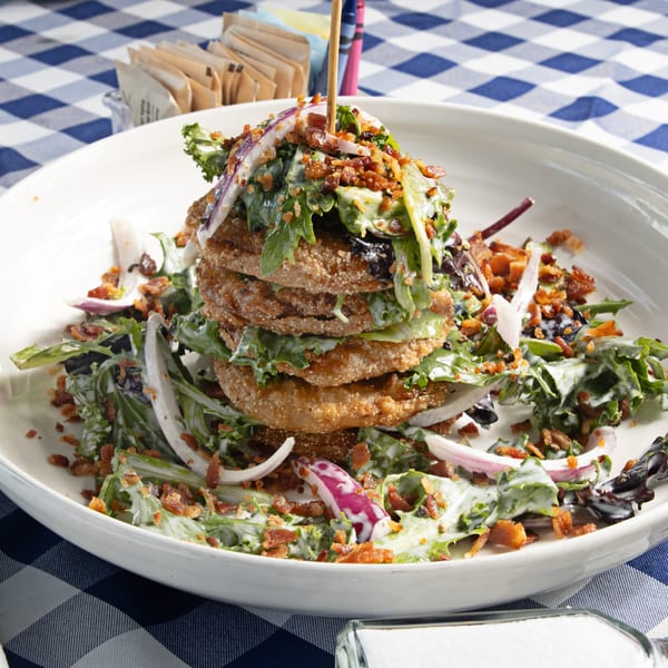 Stacked fried green tomatoes with lettuce, red onion, bacon bits, and dressing on a white plate.