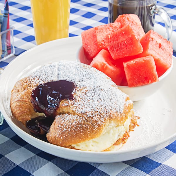 A croissant with chocolate and powdered sugar, scrambled eggs, and watermelon on a blue checkered tablecloth.