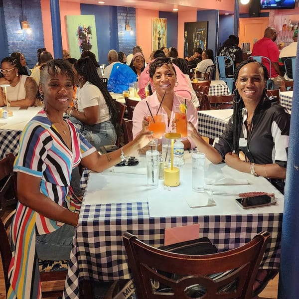 Three women at The Breakfast Boys restaurant toast with orange drinks at a checkered table.