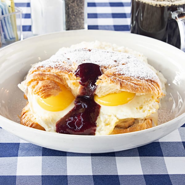 Croissant with fried eggs, jam, and powdered sugar in a white bowl on a blue and white checkered tablecloth.