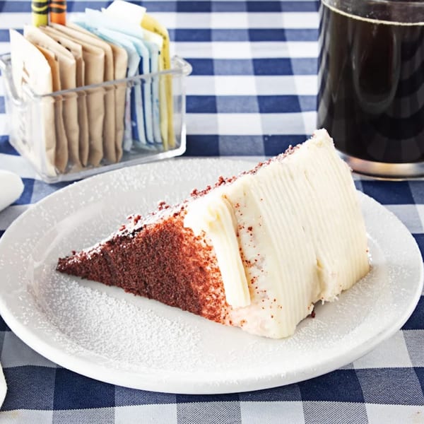 A slice of red velvet cake with white frosting and powdered sugar on a white plate.